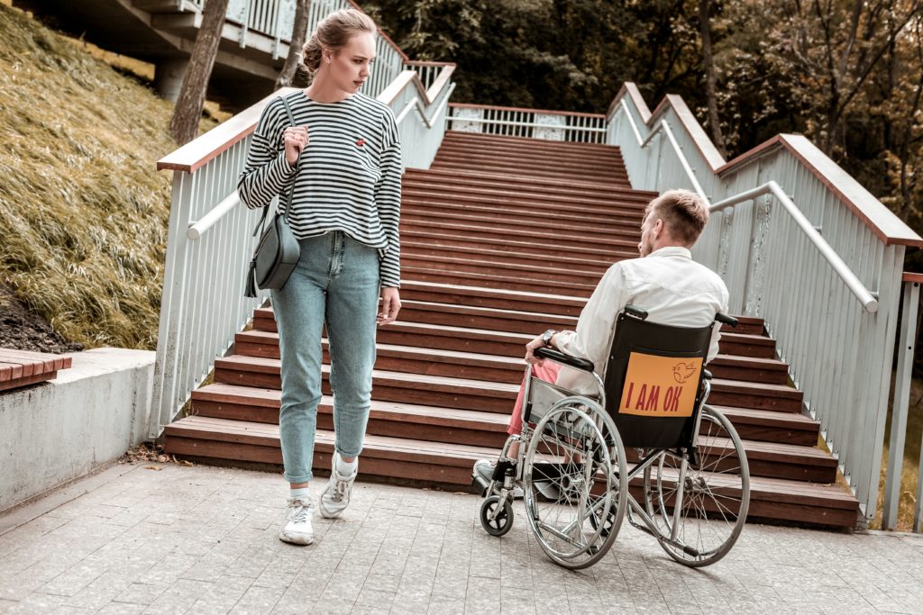Mann im Rollstuhl steht vor einer Treppe, eine Frau sieht ihn beim Vorbeigehen komisch an