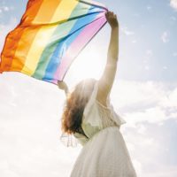 Frau in wei&szlig;em Kleid streckt eine wehende Pride-Flagge Richtung Himmel
